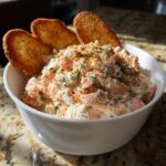 Close-up of creamy smoked salmon dip topped with herbs and spices, served with three toasted crackers.