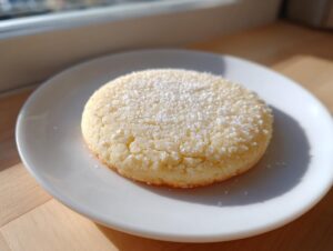 A single, soft, round sugar cookie sprinkled generously with coarse sugar, sitting on a white plate.