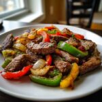 Close-up of perfectly cooked steak fajitas mixed with sautéed red, green, and yellow bell peppers and onions on a white plate.