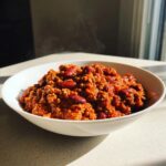 A steaming white bowl filled with rich, red high protein chili featuring ground meat and kidney beans.