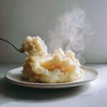 A fork lifts a scoop of steaming hot slow cooker mashed potatoes from a mound on a white plate.