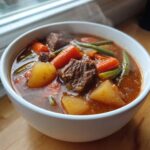 Close-up of a steaming white bowl filled with rich vegetable beef soup, featuring chunks of beef, potatoes, carrots, and green beans.