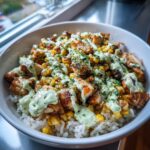A close-up of a street corn chicken rice bowl topped with grilled chicken, roasted corn, and a drizzle of creamy green sauce.