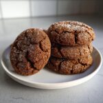 A stack of four rich, dark brown gingersnap cookies heavily coated in sparkling white sugar.