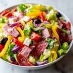 A vibrant, close-up view of a hearty grinder salad featuring lettuce, salami, ham, cheese cubes, peppers, and red onion.