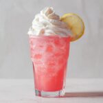 A tall glass of fizzy pink lemonade topped with whipped cream and a lemon slice.
