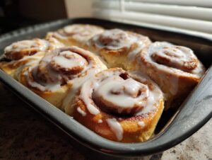 Close-up of six golden brown cinnamon rolls with thick white icing in a baking pan, made using heavy cream.