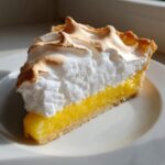 A perfect slice of lemon meringue pie showing thick, bright yellow curd and a tall, fluffy, lightly browned meringue topping.