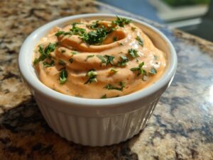 Close-up of creamy, orange-hued remoulade sauce topped with fresh chopped parsley in a white dish.