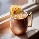 A frosty copper mug filled with an apple cider moscow mule cocktail, garnished with thin apple slices.