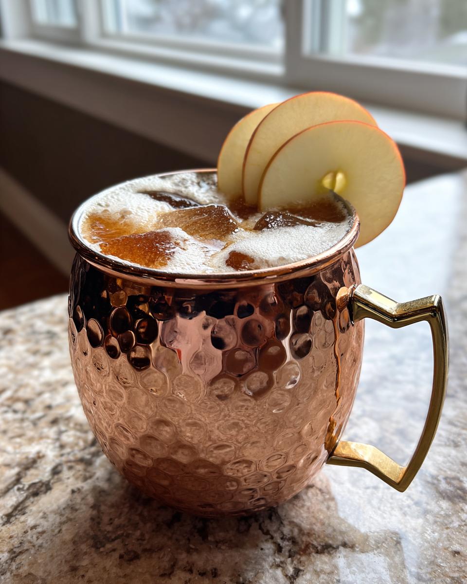 A close-up of an apple cider moscow mule cocktail served in a hammered copper mug, garnished with apple slices.