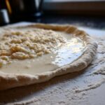 Close-up of raw pizza dough being topped with creamy white pizza sauce and crumbled cheese.