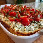 Close-up of a baked bruschetta dip featuring creamy cheese base topped with halved cherry tomatoes and fresh basil.
