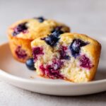A blueberry protein muffin cut in half showing the moist, berry-filled interior on a light plate.