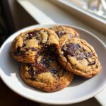Three gooey brown butter chocolate chip cookies topped with flaky sea salt on a white plate.