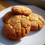 Close-up of four golden brown butter sugar cookies stacked slightly on a white plate, topped with coarse sugar crystals.