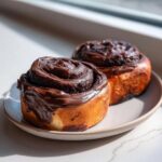 Close-up of two freshly baked chocolate cinnamon rolls generously topped with thick, glossy chocolate frosting, sitting on a plate.