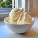 Three generous scoops of creamy banana ice cream served in a white bowl near a bright window.