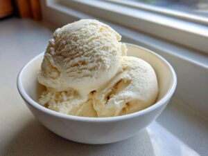 Two scoops of creamy, pale yellow banana ice cream served in a small white bowl near a window.