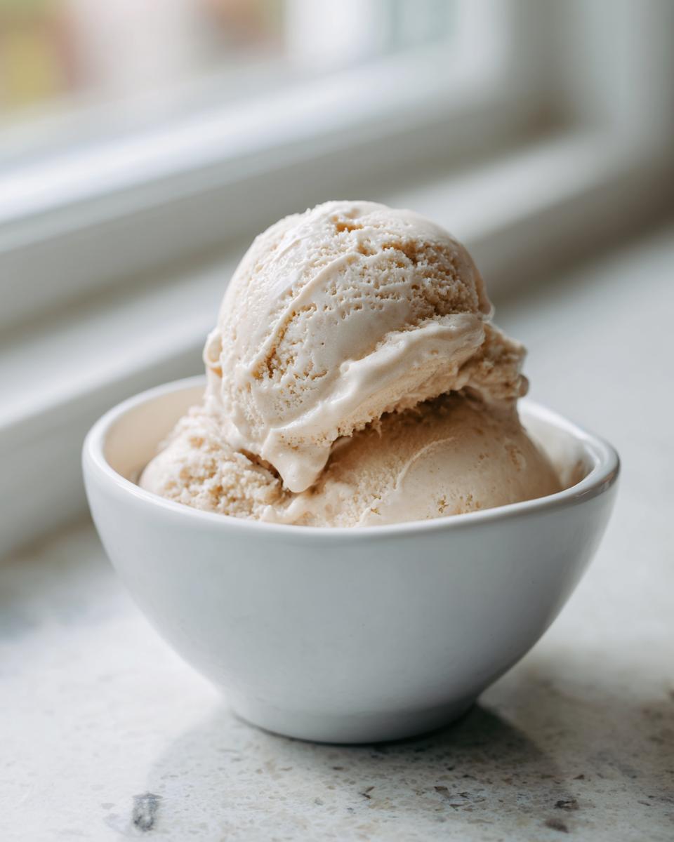 Two scoops of creamy, light brown banana ice cream served in a small white bowl near a bright window.