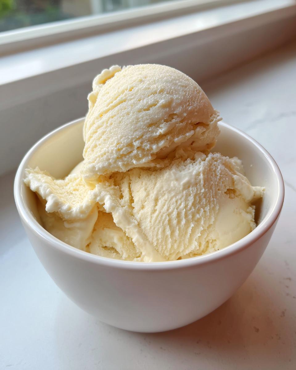 Two scoops of creamy, pale yellow banana ice cream served in a white bowl near a bright window.