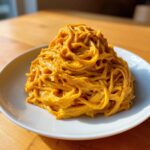 A mound of spaghetti coated in a rich, orange butternut squash pasta sauce served on a white plate.