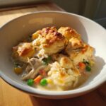 A close-up of a bowl of creamy chicken and dumplings with biscuits, featuring shredded chicken and mixed vegetables.