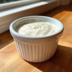 A small white ramekin filled with creamy horseradish sauce, speckled with pepper, sitting on a wooden surface.