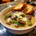 A close-up of a white bowl filled with creamy philly cheese steak soup, topped with beef chunks, green peppers, melted cheese, and two toasted bread slices.