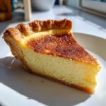 A close-up slice of rich, creamy custard pie with a deeply browned top, sitting on a white plate.