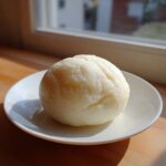 A single, smooth, pale yellow ball of fufu resting on a small white plate, illuminated by natural light.
