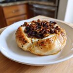 A golden puff pastry wrapped baked brie topped with caramelized pecans and dried fruit, served on a white plate.