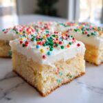 A close-up of a square sugar cookie bars slice topped with thick white frosting and colorful round sprinkles.
