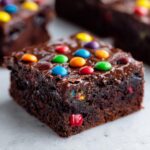 A close-up of a fudgy cosmic brownie square topped with shiny chocolate glaze and colorful candy-coated chocolates.