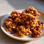 Close-up of crunchy clusters of homemade gingerbread granola mixed with dried cranberries on a light plate.