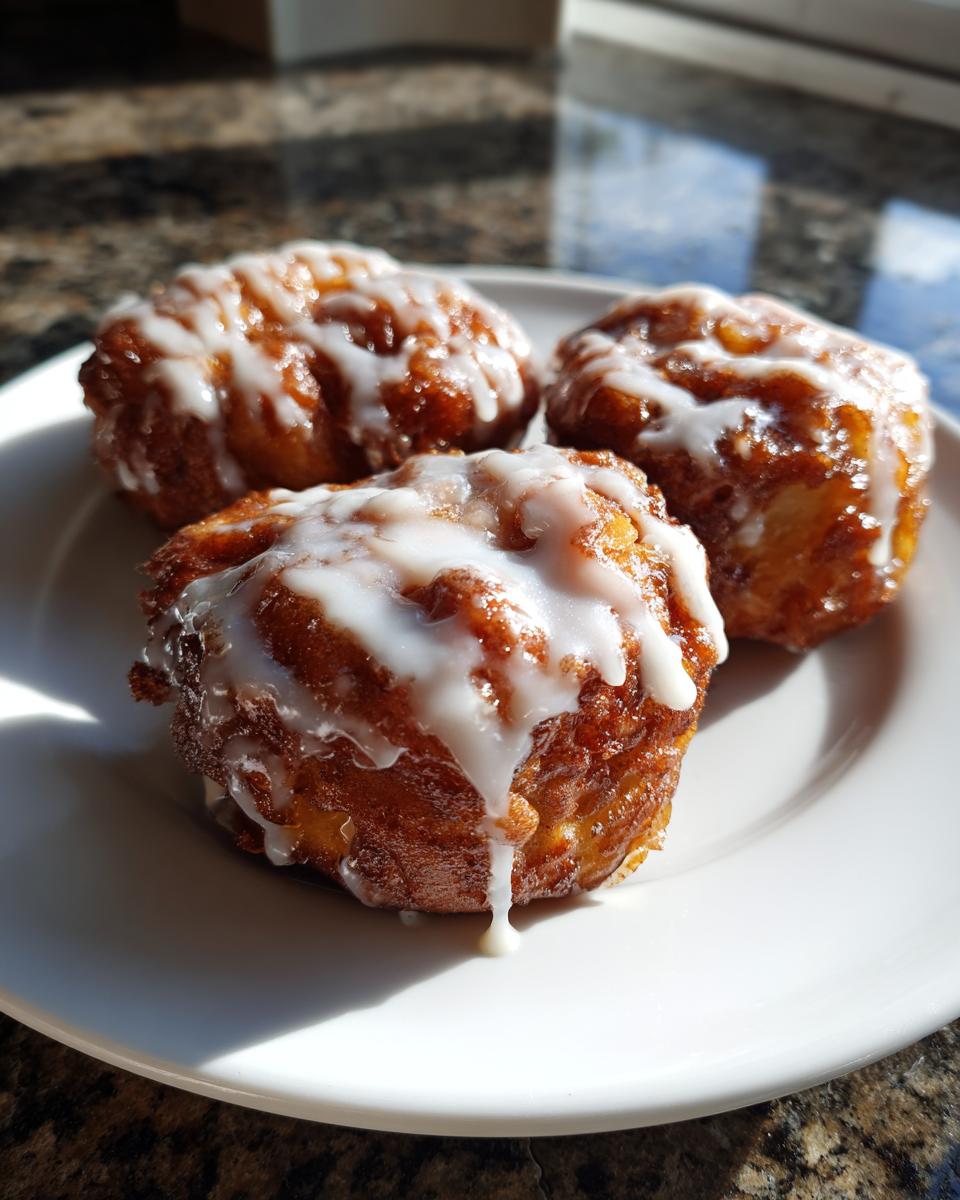 Three golden brown, irregularly shaped apple fritter pastries drizzled generously with white glaze, sitting on a white plate.