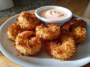 A plate of perfectly golden and crispy coconut shrimp recipe served with a small bowl of dipping sauce.
