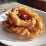 Close-up of two golden brown, crispy buñuelos generously coated in cinnamon sugar, resting on a white plate.