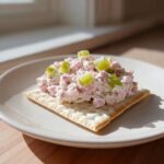 A scoop of creamy ham salad recipe topped with diced celery served on a square cracker on a white plate.