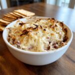 A close-up of a baked, hot reuben dip topped with melted, slightly browned cheese, served with crackers.