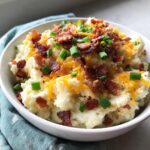 A close-up of creamy loaded mashed potatoes topped generously with crispy bacon bits, shredded cheddar cheese, and chopped green onions.