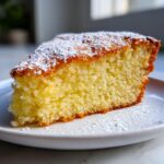 Close-up of a moist slice of olive oil cake dusted with powdered sugar on a white plate.