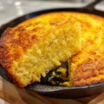A close-up of a moist slice of golden sourdough cornbread being lifted from the rest of the loaf in a black cast iron skillet.