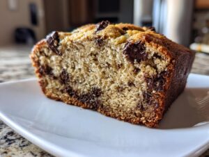 Close-up of a moist slice of vegan banana bread packed with melted chocolate chips, resting on a white plate.
