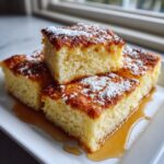 Four square pieces of fluffy sheet pan pancakes stacked and drizzled with syrup and powdered sugar.