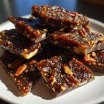 Close-up of shiny, dark brown Pecan Pie Bark pieces stacked on a white plate, loaded with pecans.