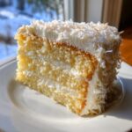 A close-up slice of a moist, three-layer coconut cake recipe covered in white frosting and shredded coconut.