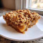 A close-up of a square slice of warm pear crisp with a thick, golden oat crumble topping on a white plate.