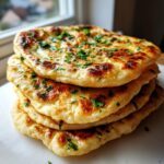 A stack of four freshly made, golden brown naan bread pieces brushed with butter and topped with chopped parsley.