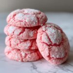 A stack of four bright pink, crinkle-topped strawberry cake mix cookies dusted heavily with powdered sugar.